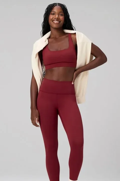 Frau in bordeauxroter Leggings mit passendem Sport-BH und heller Strickjacke über der Schulter, von Kopf bis Wade