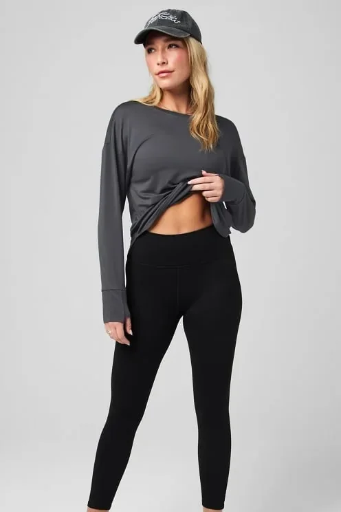 Frau in schwarzer Leggings und grauem Longsleeve mit Kappe, von Kopf bis Knöchel vor hellgrauem Hintergrund