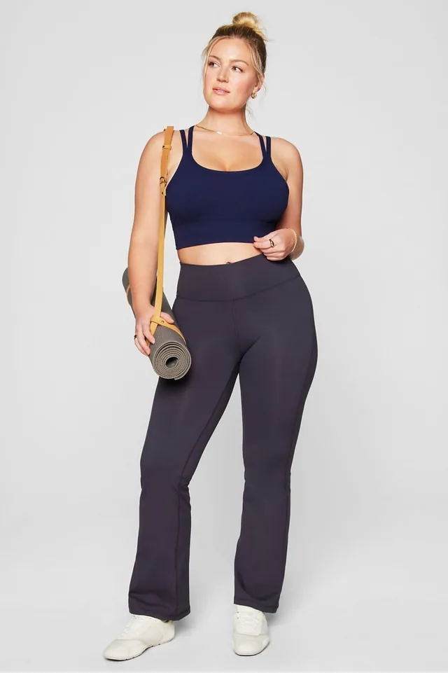 Frau in dunkelblauem Sport-Top und dunkler Leggings, eine Yogamatte unter dem Arm